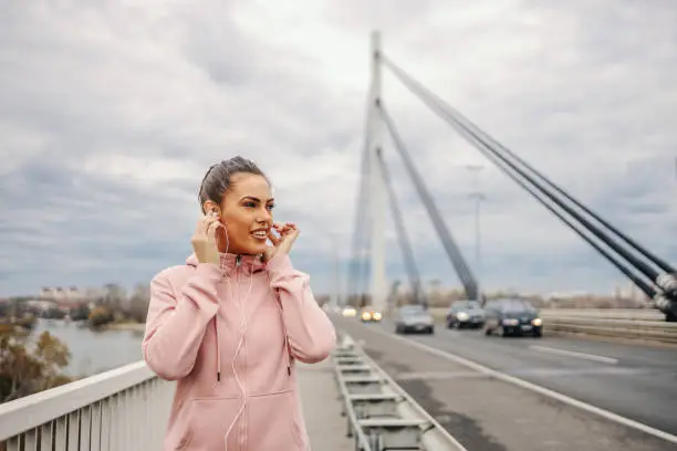 Photo of Beautiful young woman in sportswear standing on the bridge, putting earphones and preparing for jogging. Outdoor fitness on cloudy weather concept.