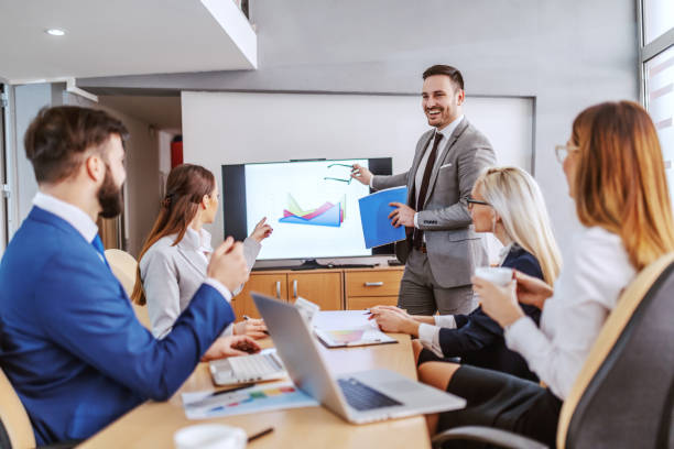 istock Young handsome caucasian bearded chief standing in boardroom, holding paperwork and having presentation. Other colleagues listening to him and and asking him questions. 1198699795