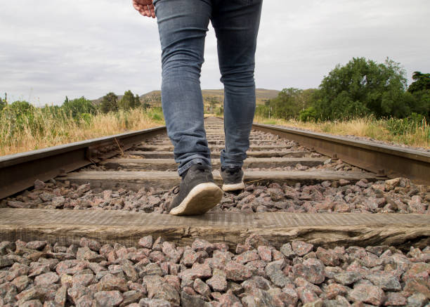 piedi giovani irriconoscibili che camminano sui binari della ferrovia in una scena rurale in una giornata nuvolosa - railroad track train journey rural scene foto e immagini stock