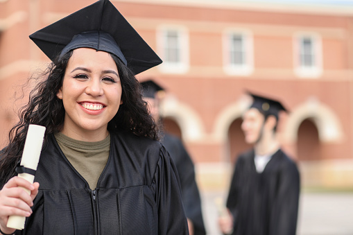 Latin descent girl holds diploma after high school or college graduation ceremony.   She looks at camera with a big smile as she wears a black cap and gown.