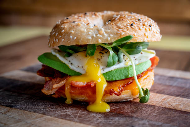 colazione bagel con uovo, verdure, avocado, pancetta, salsiccia e formaggio su un taglio rustico - sandwich eggs bacon breakfast foto e immagini stock