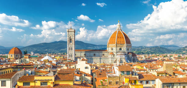 Top aerial panoramic view of Florence city with Duomo Cattedrale di Santa Maria del Fiore cathedral, buildings houses with orange red tiled roofs and hills range, blue sky white clouds, Tuscany, Italy vista panorámica aérea superior de la ciudad de florencia con la catedral duomo cattedrale di santa maria del fiore, casas de edificios con techos de tejas rojas anaranjadas y la cordillera de las colinas, nubes blancas cielo azul, toscana, italia