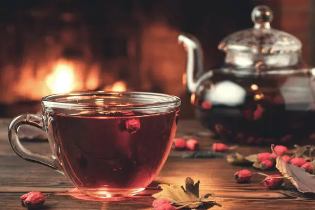 Photo of Tea with hawthorn in a glass cup and teapot on a wooden table in a room with a burning fireplace, closeup