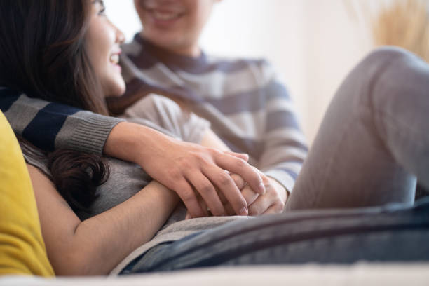 Lovely Asian couple sitting on bed in bedroom talking together with happiness. Boy hugging girl and holding hand, be hand in hand. Crop shot focus on hands. Man looking at woman eyes and smile face. Lovely Asian couple sitting on bed in bedroom talking together with happiness. Boy hugging girl and holding hand, be hand in hand. Crop shot focus on hands. Man looking at woman eyes and smile face. couple holding hands stock pictures, royalty-free photos & images