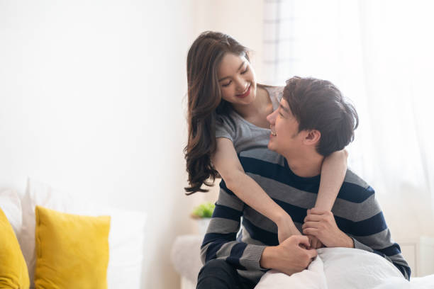 homem e mulher asiáticos dos pares novos que permanecem junto no quarto de cama. menina que abraça o menino de atrás, olhando os olhos em se sentindo o amor feliz com cara do sorriso. relacionamento do conceito novo da menina e do menino - japanese ethnicity family smiling happiness - fotografias e filmes do acervo