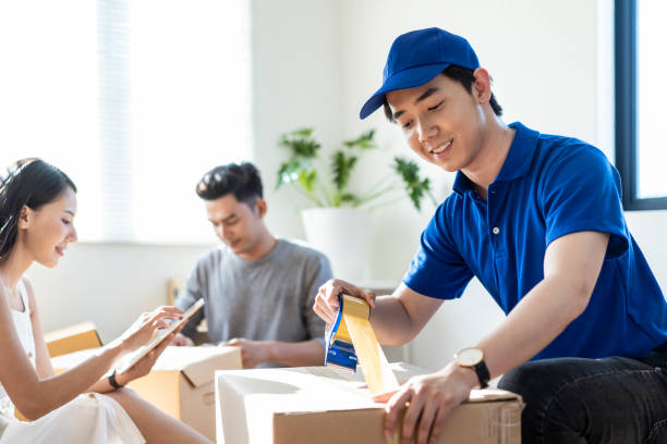 House moving and packing service. Asian man in blue shirt uniform using tape to package box at customer house. Customer couples check things before boxing. Home delivery and moving service concept. House moving and packing service. Asian man in blue shirt uniform using tape to package box at customer house. Customer couples check things before boxing. Home delivery and moving service concept. Hiring Movers stock pictures, royalty-free photos & images