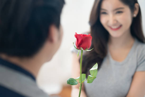 かわいい女性に赤いバラの花を与えるアジアの若者。素敵なバレンタインプレゼントを受け取る女の子は、愛を感じ、顔に恥ずかしがり屋の笑顔。美しい花に焦点を当てます。男の子と女の� - engagement ストックフォトと画像