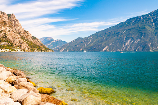 Round boulders lying on the shore of beautiful Garda lake in Lombardy, Italy surrounded by high dolomite mountains. Classic white sailing yacht floating on the lake