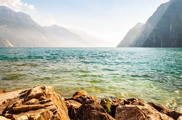 Photo of Rocky stones lying on the shore of beautiful Garda lake in Lombardy, Italy surrounded by high dolomite mountains. Sunbeams penetrating from above the rocks and warming misty fog above the water