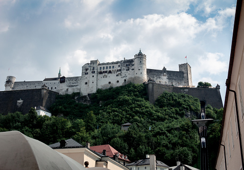 View of Hohensalzburg fortress from below with modern funicular trains going up and down. Hill covered in green bushes, trees and railroad. Old town Salzburg