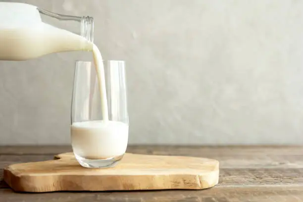Kefir, milk or Turkish Ayran drink are poured into a glass cup from a bottle. A glass stands on a wooden stand on a rustic wooden table. Place for text.