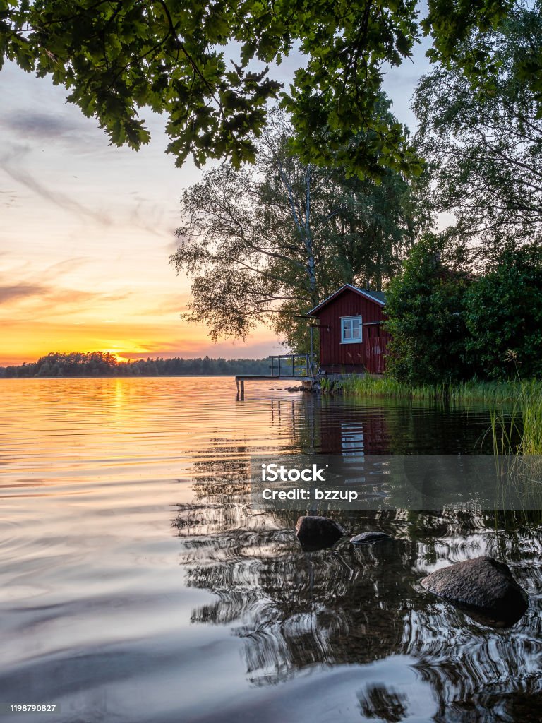 Bild av solnedgång vid sjön med träd, skandinaviskt trähus och blå himmel - Royaltyfri Sommar Bildbanksbilder