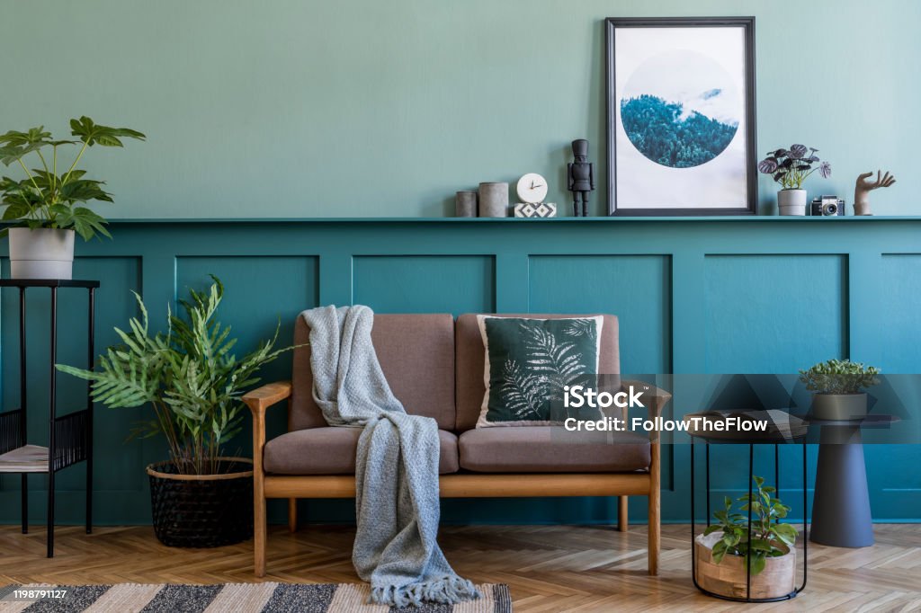 Stylish interior design of living room at apartment with brown sofa, a lot of plants, design furnitures and elegant accessories. Mock up poster frames on the shelf. Green wood panelling. Home staging. Stylish interior of living room with design furniture. Green wood panelling with shelf. Modern home decor. Mock up poster frame. Template. Home Interior Stock Photo
