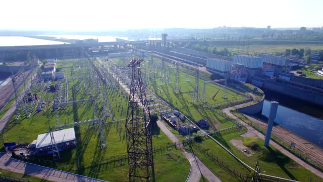 An aerial circular orbit view a high voltage electric power tower, hydroelectric power station and electric substation with tall pylons and hog voltage distribution cables. Morning spring summer sun.