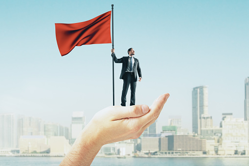 Abstract hand holding businessman with red flag on blurry New York city background. Leadership and success concept