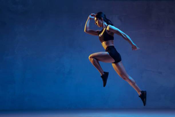 Caucasian young female athlete practicing on blue studio background in neon light Caucasian young female athlete practicing on blue studio background in neon light. Close up of sportive model jumping high, running. Body building, healthy lifestyle, beauty and action concept. strength training photos stock pictures, royalty-free photos & images