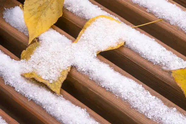 Wooden bench seat closeup. Covered with snow and yellow autumn leaves. Selective focus. Winter outdoor scene.