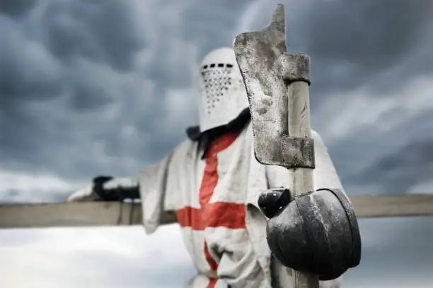 Side view of medieval knight with red cross on armor posing and holding axe. Selective focus of weapon in crusader arms standing outdoors and leaning on handrail. Concept of warrior.
