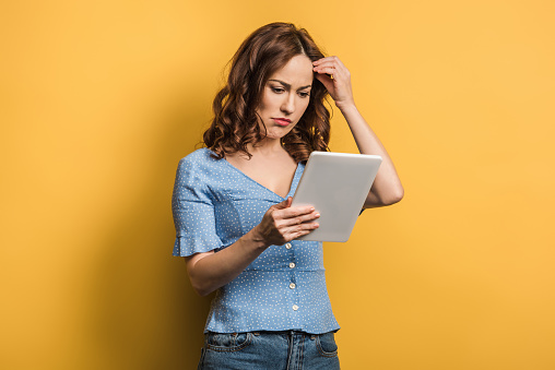thoughtful woman touching head while using digital tablet on yellow background