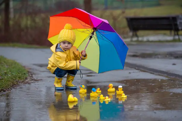 Photo of Beautiful funny blonde toddler boy with rubber ducks and colorful umbrella, jumping in puddles and playing in the rain