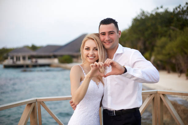 noiva e noivo felizes que sorriem e que mostram o sinal do coração com suas mãos em uma praia tropical do recurso luxuoso dos termas em maldives. casamento e lua de mel na ilha tropical - honeymoon wedding beach couple - fotografias e filmes do acervo