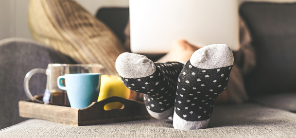 Woman sitting on a couch in the living room with warm socks in a winter morning. Girl using laptop and works at home, having natural breakfast with tea coffee and fruits. Focus on feet in foreground