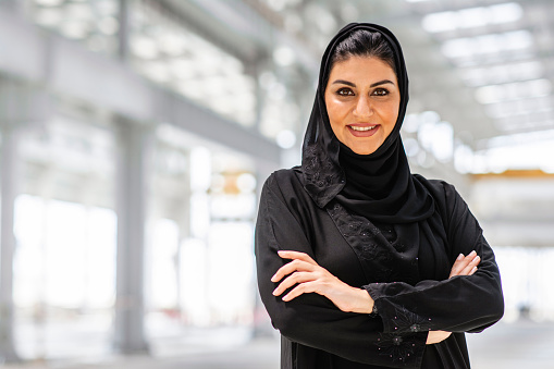 Portrait of smiling Abu Dhabi female design professional wearing traditional Islamic clothing and smiling at camera with arms crossed at construction site.