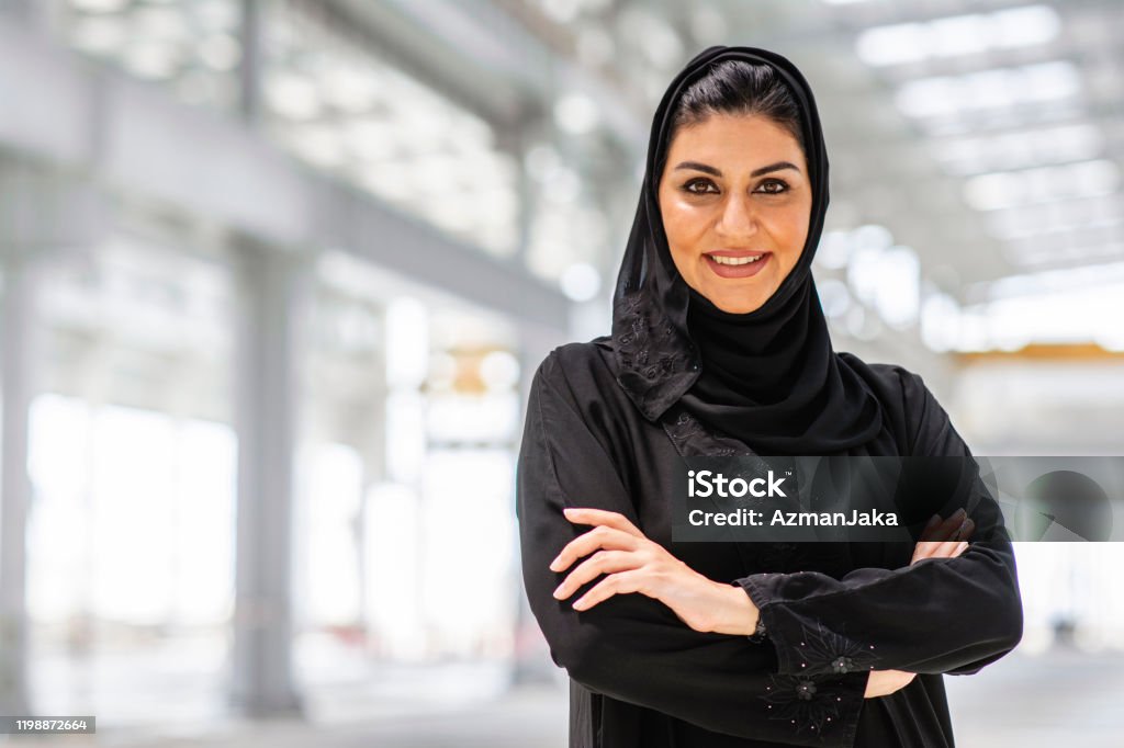 Confident Middle Eastern Female Construction Professional Portrait of smiling Abu Dhabi female design professional wearing traditional Islamic clothing and smiling at camera with arms crossed at construction site. One Woman Only Stock Photo