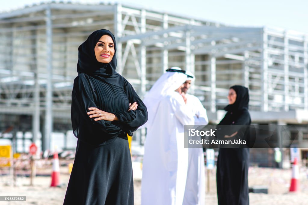 Abu Dhabi Female Construction Professional and Colleagues Confident Middle Eastern female design professional in traditional Islamic clothing looking at camera with arms crossed and coworkers in background. United Arab Emirates Stock Photo
