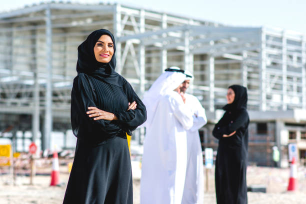 Abu Dhabi Female Construction Professional and Colleagues Confident Middle Eastern female design professional in traditional Islamic clothing looking at camera with arms crossed and coworkers in background. emirati culture photos stock pictures, royalty-free photos & images