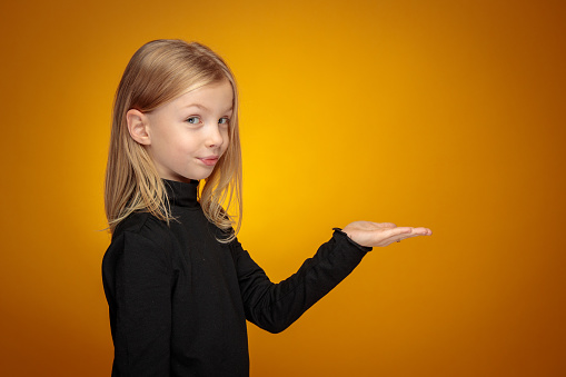 A portrait of a cheerful little blonde girl holding hand pulm up in front of her and looking at the camera. Studio shot on yellow background.