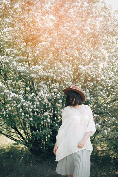 istock Happy boho woman in hat having fun in sunny light near white blooming tree in spring park. Stylish hipster girl enjoying spring and dancing. Atmospheric moment of happiness 1198884801