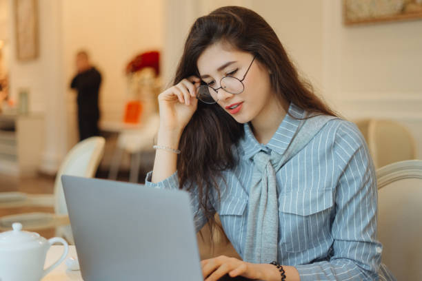 istock Cute asian woman having a cup of tea while using laptop computer in cafe and touching her glasses 1198907077