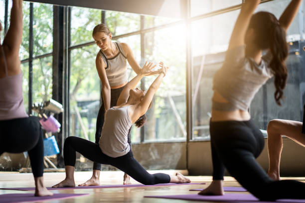 grupo de povos multi étnicos que aprendem a classe da ioga no clube da aptidão. o instrutor caucasiano fêmea que treina e ajusta o pose correto ao estudante asiático da menina na parte dianteira quando outro os seguir. - healthy lifestyle yoga vitality flexibility - fotografias e filmes do acervo