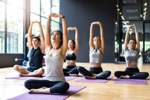 groupe de personnes multi éthique apprenant le cours de yoga dans le club de forme physique. l'instructeur asiatique féminin menant meditate s'asseyant la main et la pose de bras, les étudiants suivent derrière. yoga pour la santé, concept de bien-êt - yoga class caucasian young adult group of people photos et images de collection