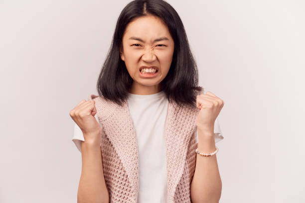 human looses temper, clenches teeth wrinkles nose Portrait of irritated human looses temper, clenches teeth from anger, wrinkles nose, looks with hate at camera. Woman Asian appearance brown eyes stands isolated white background in Studio clenching teeth stock pictures, royalty-free photos & images