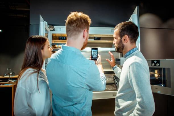 Store seller explaining the compartments usage in a new generation fridge to a young couple in a kitchen appliances store