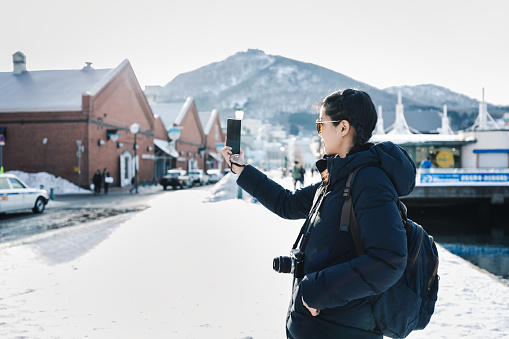 Winter snow travel concept, Happy traveler asian woman with backpack and camera using mobile phone to selfie in red brick warehouse at Hakodate city, Hokkaido, Japan
