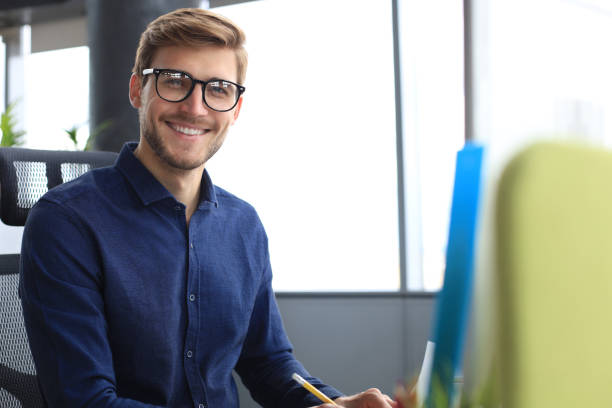 istock Good looking young business man looking at camera and smiling while sitting in the office. 1198989840