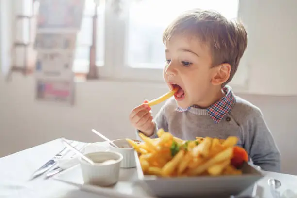 Photo of Portrait of small little cute caucasian boy kid eating french fries potato chips at the table in the restaurant or at home three or four years old