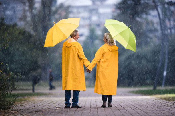 back view of happy senior couple with umbrellas talking on rainy day. - umbrella senior adult couple autumn imagens e fotografias de stock