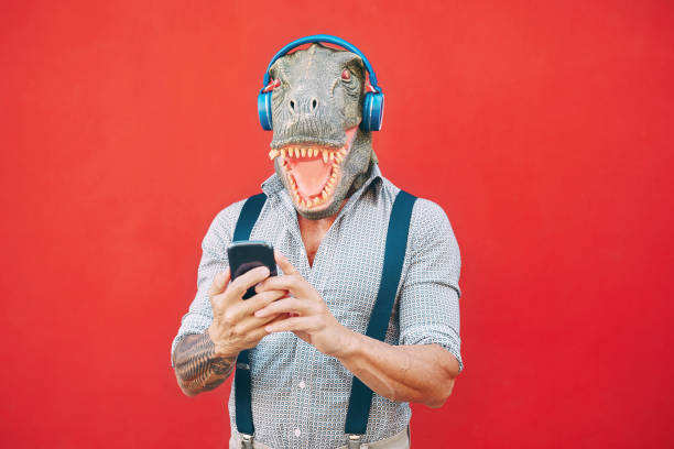 senior fashion man wearing t-rex mask using mobile smartphone listening music with headphones - crazy bizarre hipster guy having fun with new technology - funny and absurd surreal concepts senior fashion man wearing t-rex mask using mobile smartphone liste - headphones music mobile phone tattoo zdjęcia i obrazy z banku zdjęć