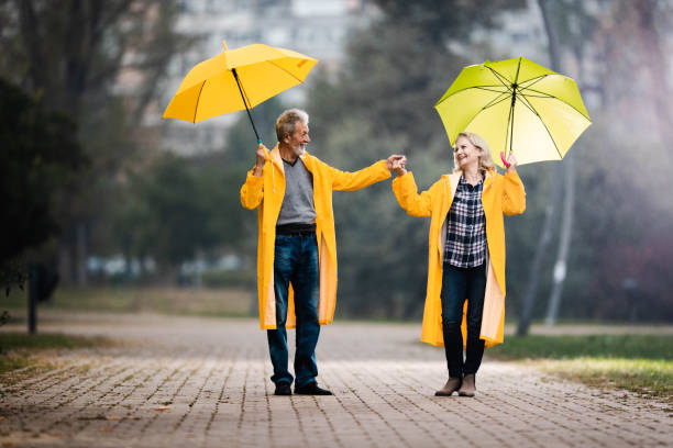 couples mûrs heureux dans les imperméables jaunes marchant avec des parapluies un jour pluvieux. - umbrella senior adult couple autumn photos et images de collection