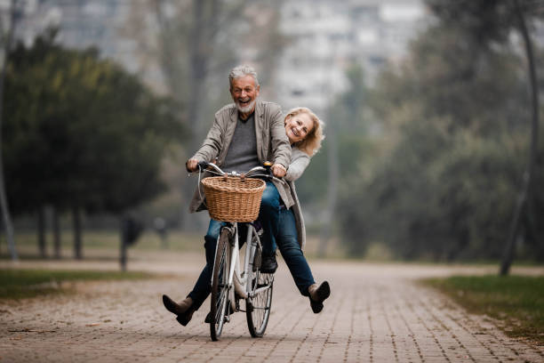coppia senior giocosa che si diverte in bicicletta nella giornata autunnale. - autumn women park people foto e immagini stock