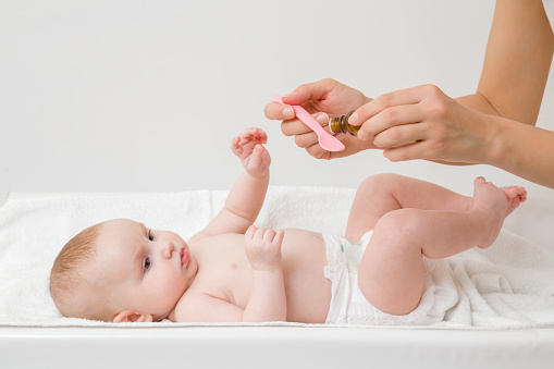 Young mother hands holding pink plastic spoon and bottle of medicines. Infant lying down on changing table and receiving vitamins. Side view. Close up.