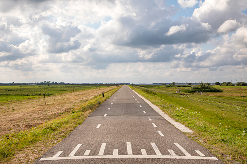 Road in Holland with white cycle path lines on both sides, perspective, under heavy threatening cloudy skies and between green meadows and a faraway straight horizon.