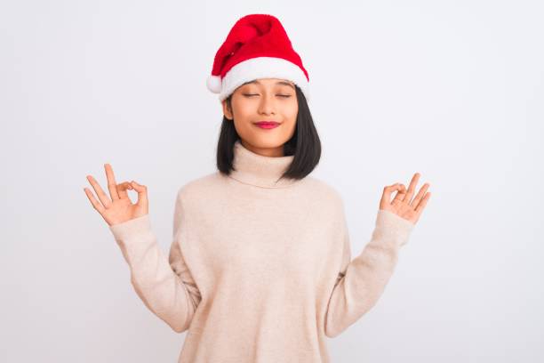 jeune belle femme chinoise utilisant le chapeau de santa de noel au-dessus du fond blanc dissourssaré détendent et souriant avec des yeux fermés faisant le geste de méditation avec des doigts. concept de yoga. - holiday emotional stress christmas santa claus photos et images de collection