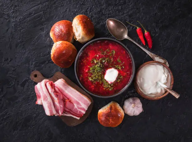 Traditional Ukrainian or Russian red vegetable and meat soup borscht in a cast-iron pot with garlic buns and sour cream on a black background. Traditional Slavic dish with beets, cabbage, tomatoes. The view from the top.