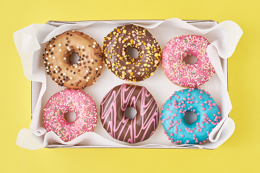 Different types of colorful donats decorated sprinkles and icing in box on yellow background, top view