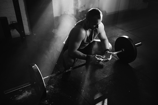 Black and white photo of muscular build man using sports chalk on hands while weightlifting in a gym.